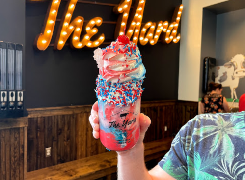 A colorful dessert drink topped with whipped cream, sprinkles, and a cherry, held in front of a sign that says "The Yard."