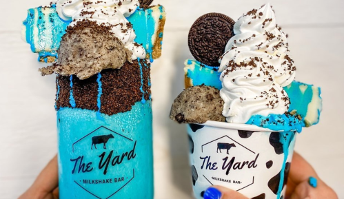 Two colorful milkshakes topped with whipped cream, cookies, and sprinkles, featuring The Yard Milkshake Bar branding.