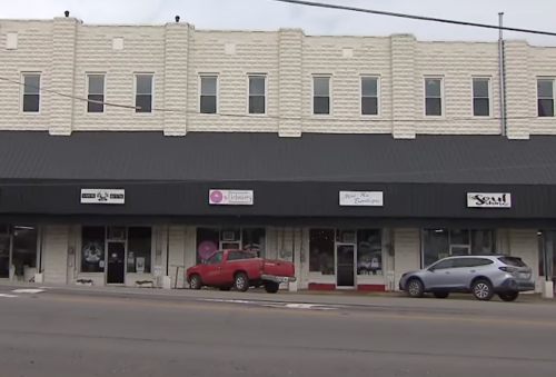 A row of storefronts with various signs, featuring a red truck parked in front and a gray car nearby.