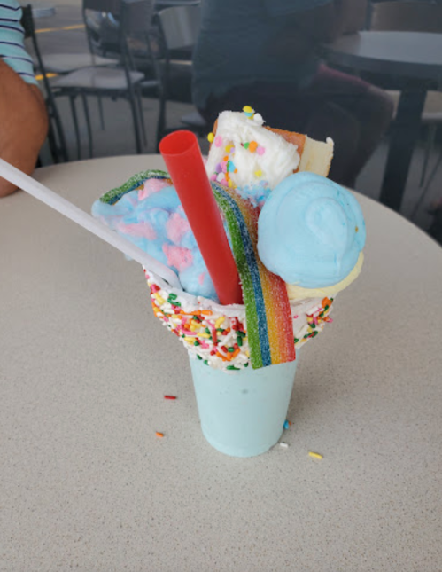 A colorful ice cream treat topped with sprinkles, cotton candy, a rainbow candy strip, and a blue scoop, served in a cup.
