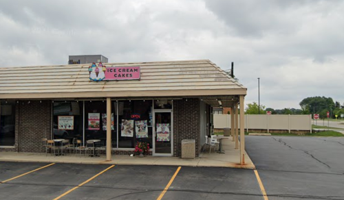 Exterior of an ice cream shop with a sign reading "Ice Cream Cakes," featuring outdoor seating and cloudy skies.