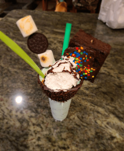 A colorful dessert with whipped cream, chocolate, sprinkles, a brownie, marshmallows, and a cookie on top.