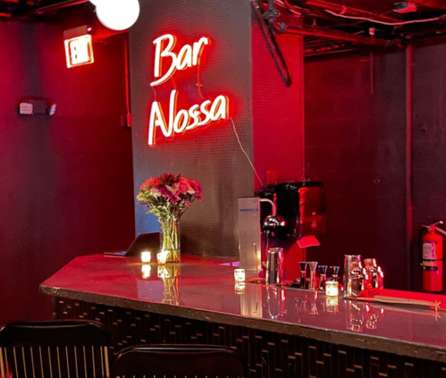 A dimly lit bar with a neon sign reading "Bar Nossa," flowers on the counter, and candles illuminating the space.