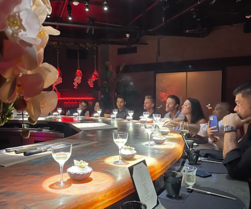 A dimly lit restaurant bar with guests seated, enjoying drinks and small dishes, surrounded by elegant decor.