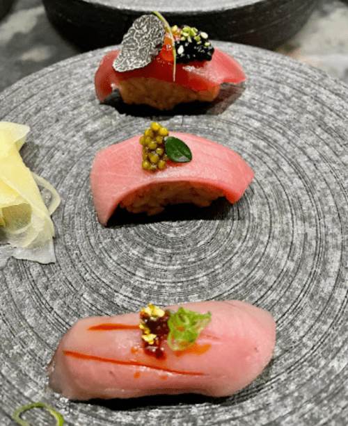 Three pieces of sushi on a textured plate, featuring tuna, caviar, and garnishes, with pickled ginger on the side.