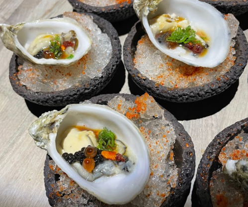 Five oysters on a bed of ice, garnished with caviar and colorful toppings, served in black stone dishes.