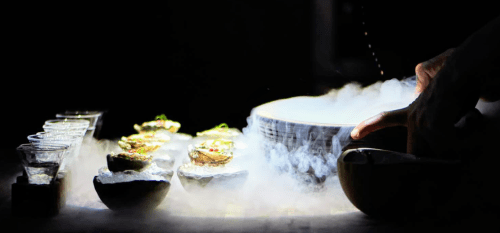 A hand prepares a dish with smoke, surrounded by elegant appetizers and shot glasses on a dark background.