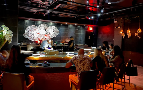 A stylish restaurant interior with a bar, patrons seated, and a chef preparing food in a vibrant atmosphere.