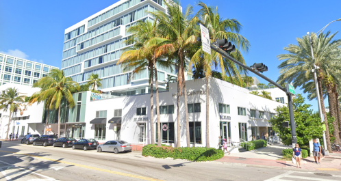 A modern building with palm trees, shops, and pedestrians on a sunny street.