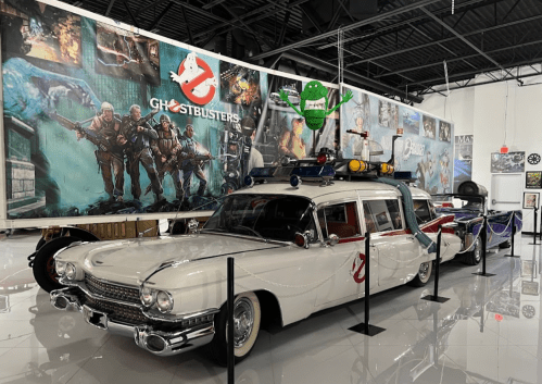A vintage Ghostbusters car displayed in a museum, with themed artwork and memorabilia in the background.