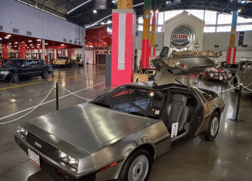 A DeLorean car with gullwing doors displayed in a vintage car museum, surrounded by other classic vehicles.