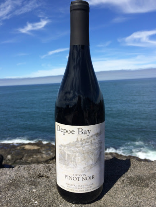 A bottle of Depoe Bay Oregon Pinot Noir stands on a stone ledge with a scenic ocean view in the background.