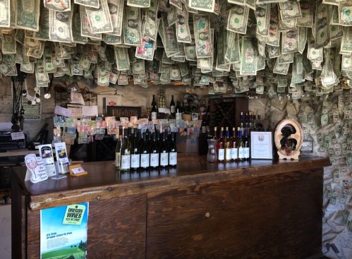 A rustic bar with bottles of wine on display, surrounded by walls covered in hanging dollar bills.