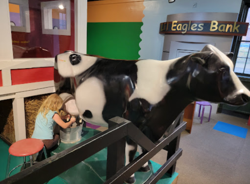 A child milks a life-sized cow model in a colorful play area designed for interactive learning.