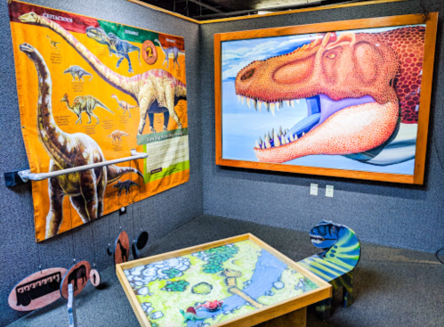 A colorful dinosaur-themed exhibit featuring a wall poster, a large dinosaur painting, and a playful table.