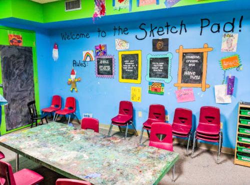Colorful classroom with a "Welcome to the Sketch Pad!" sign, a messy table, and red chairs against a blue wall.