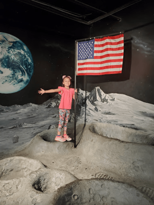 A girl stands on a moon-like surface, holding an American flag, with a painted Earth and mountains in the background.
