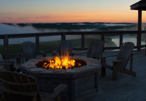 A cozy fire pit surrounded by wooden chairs, with a scenic view of fog and a sunset over the water in the background.