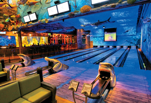 A unique bowling alley with shark-themed decor, featuring lanes, seating, and screens in an underwater ambiance.