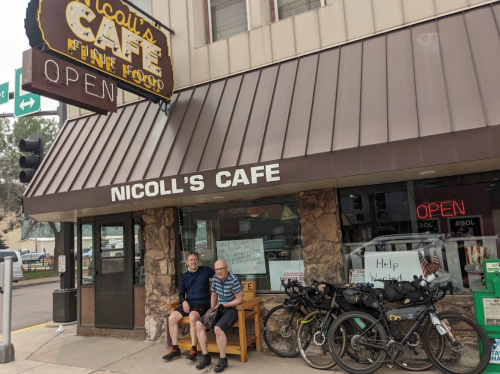 Two men sit on a bench outside Nicoll's Cafe, with bicycles nearby and a sign indicating the cafe is open.