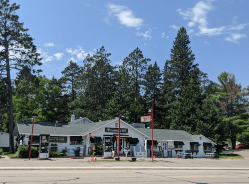 A small, rustic store with a green roof, surrounded by trees, featuring signs for ice and other goods.