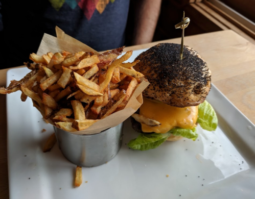 A cheeseburger with lettuce and a poppy seed bun, served with a side of crispy fries in a metal cup.
