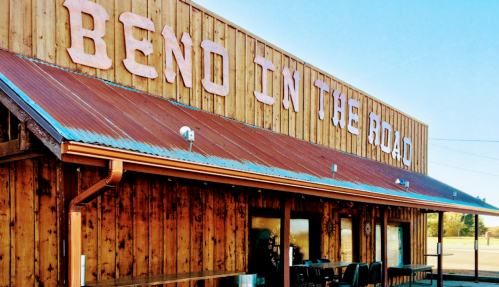 A rustic wooden building with a red metal roof, featuring the words "BEND IN THE ROAD" prominently displayed.