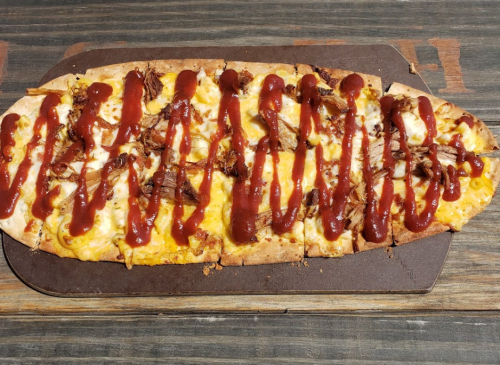 A flatbread pizza topped with cheese, pulled meat, and drizzled with barbecue sauce on a wooden surface.