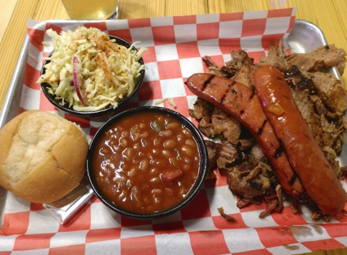 A tray with barbecue brisket, sausage, baked beans, coleslaw, and a bun on a red and white checkered paper.