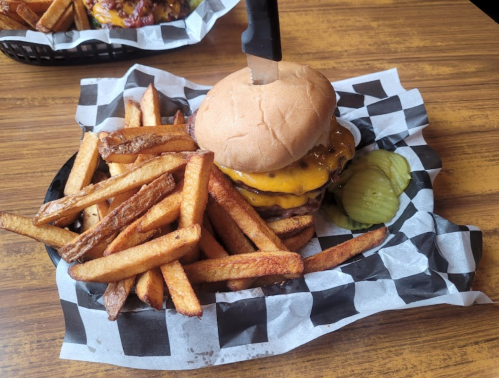 A cheeseburger with two patties, pickles, and fries on a checkered paper plate, topped with a knife.