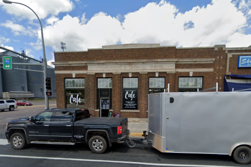 A black pickup truck towing a silver trailer in front of a historic brick bank building with a cafe sign.