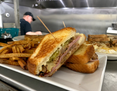 A close-up of a toasted sandwich with pickles and ham, served with fries on a white plate, in a kitchen setting.