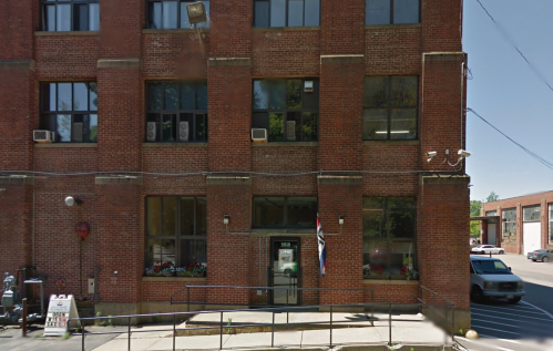 A brick building with large windows, a ramp, and flower boxes at the entrance, located on a city street.