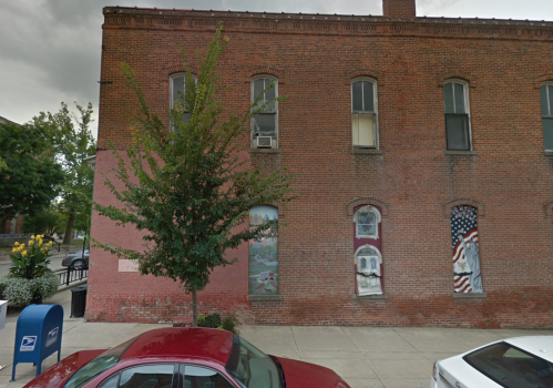 Brick building exterior with a tree, a mailbox, and murals on the wall, featuring a landscape and an American flag.