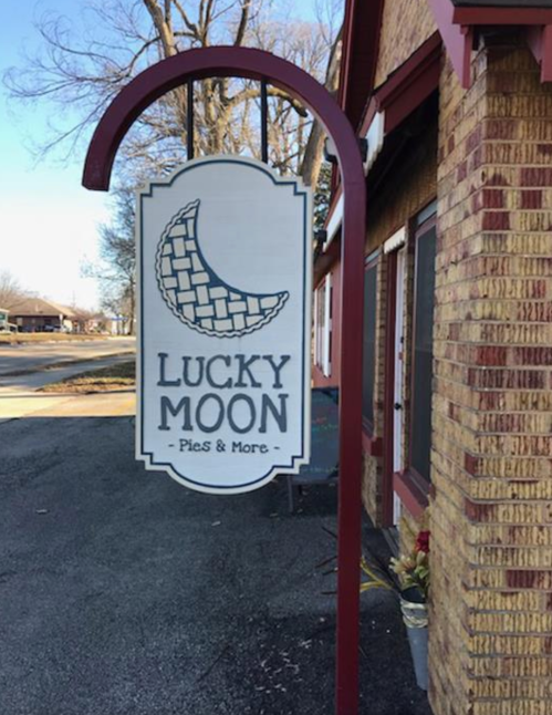 Sign for "Lucky Moon Pies & More" featuring a crescent moon design, hanging outside a brick building.