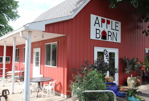 Red barn building with "Apple Barn" sign, outdoor seating, and decorative plants in front.