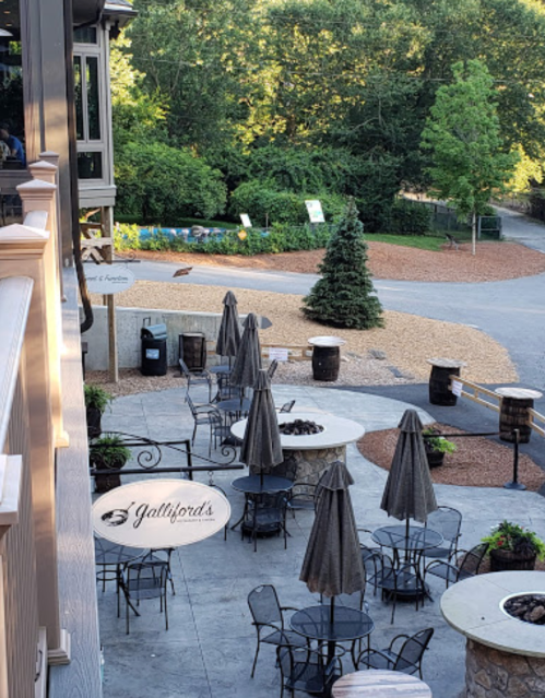 Outdoor dining area with tables, umbrellas, and a tree in a scenic setting, featuring a restaurant sign.
