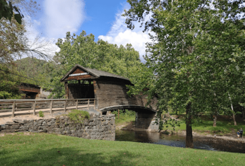 A wooden covered bridge spans a stream, surrounded by lush green trees and a clear blue sky.
