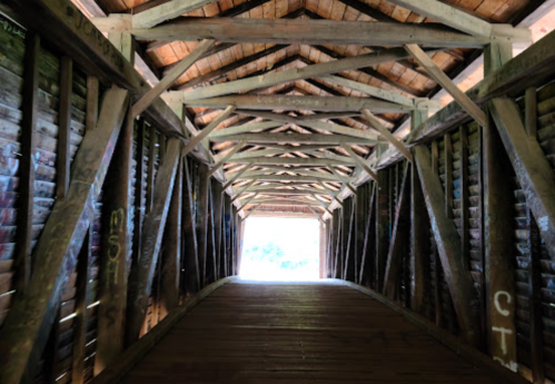 Interior view of a wooden covered bridge, showcasing beams and a bright light at the end.