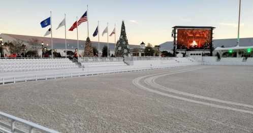 A winter scene featuring a snowy arena, empty seating, flags, and a large screen, with holiday decorations in the background.