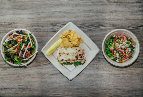 A plate with a sandwich, chips, and two bowls of colorful salads on a wooden table.