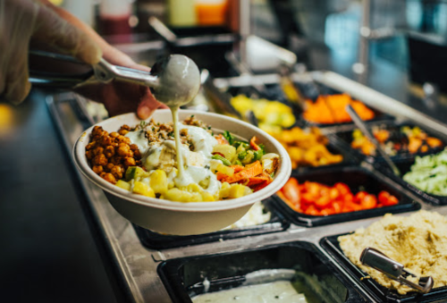 A hand scoops sauce onto a bowl filled with vegetables and grains at a salad bar with various toppings in the background.