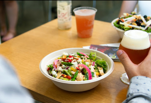 A person holds a beer while sitting at a table with a colorful salad and drinks in the background.