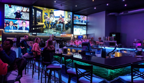 A lively bar scene with multiple screens showing sports, colorful lighting, and patrons seated at the bar.