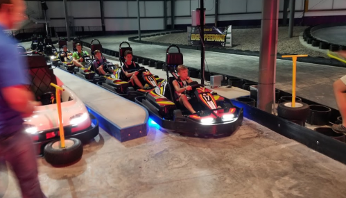 A group of kids in go-karts lined up on a track in an indoor racing facility.