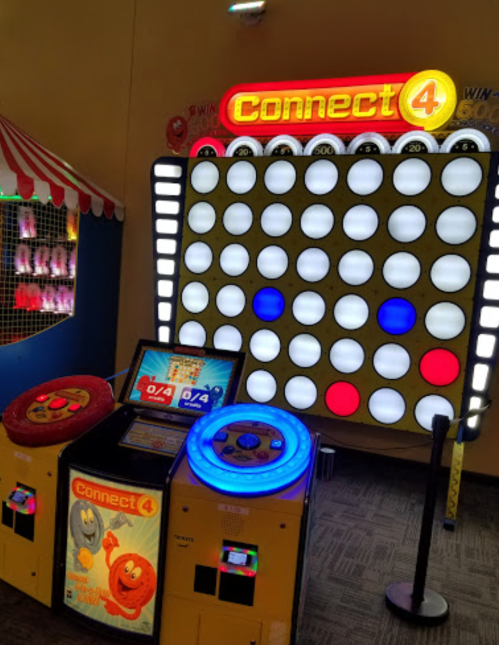 A brightly lit Connect 4 arcade game with a large board and two player stations, featuring colorful buttons and screens.