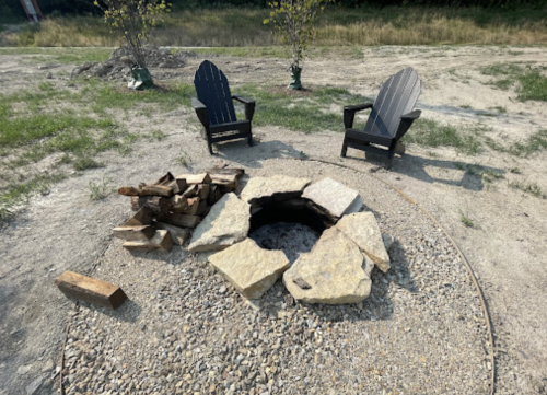 A stone fire pit surrounded by two black Adirondack chairs and a pile of firewood on a grassy area.