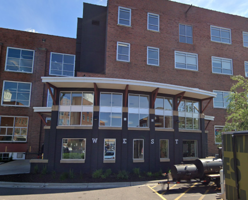 Modern brick building with large windows and a prominent entrance labeled "WEST." Outdoor area with landscaping.