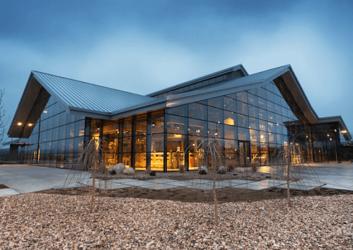 Modern glass building with a sleek design, illuminated at dusk, surrounded by landscaped gravel and plants.