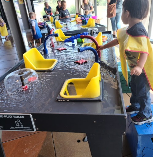 Children play at a water table with yellow buckets and colorful toys, while adults supervise nearby.